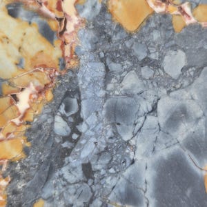 May include: Close-up of a natural stone surface with a complex pattern. The stone features a mix of colors, including shades of gray, yellow, and hints of red. The surface has a polished appearance, with visible veins and variations in texture.