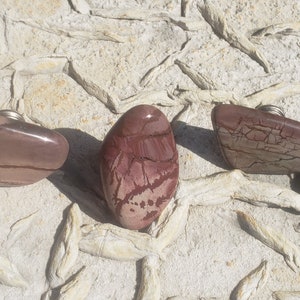 May include: Three polished stone bottle stoppers in shades of red and brown, each with a silver-colored metal base. The stones have unique patterns and shapes, resting on a textured, light-colored surface.