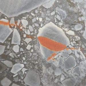May include: A close-up view of a terrazzo surface featuring a dense composition of gray, white, and off-white stone fragments embedded in a concrete base, accented by a prominent, elongated, vibrant orange vein running across the center.
