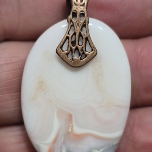 May include: Oval pendant with a white and cream-colored stone, featuring swirls of orange and gray. The pendant has a decorative bronze-colored bail. This handcrafted jewelry piece is suitable for necklaces.