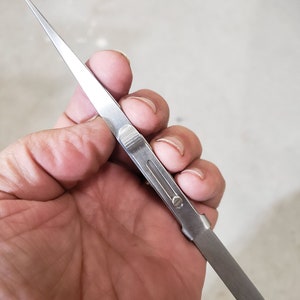 May include: A stainless steel tweezer held in a hand. The tweezer has a pointed tip with fine serrations and a flat, rectangular handle. The tweezer is designed for precision work.