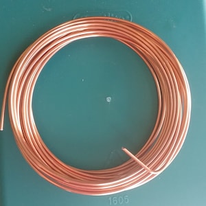 May include: A coil of copper wire, wound tightly in a circle, on a green surface. The wire is a bright, shiny copper color. The number 1605 is printed on the surface below the wire.