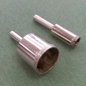 May include: Two silver diamond-tipped drill bits on a green surface. One bit is cylindrical with a larger diameter, and the other is smaller. The cutting edges of the bits are visible, showing a textured, abrasive surface.