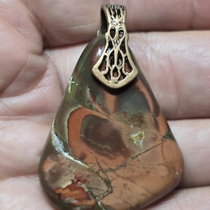 May include: A teardrop-shaped pendant with a bronze-colored bail. The stone has a mix of brown, green, and orange hues with natural patterns and cracks. The bail has an openwork design.