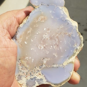 May include: A polished, irregularly shaped stone with a mix of pale blue, cream, and tan hues. The stone has a translucent quality, with visible patterns and textures. The edges are rough, and the overall appearance is natural and organic.