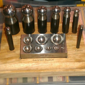 May include: A set of metal dapping tools and a dapping block. The tools have rounded tops and cylindrical bodies. The dapping block is rectangular with various sized rounded depressions. The set is on a wooden base.