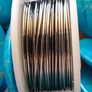 May include: A spool of silver wire wrapped around a white plastic bobbin. The wire is shiny and has a metallic finish.