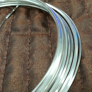 Puede incluir: Primer plano de una bobina de alambre brillante de color plateado. El alambre es liso y reflectante, con una ligera curva. El fondo es una tela marrón texturizada. El alambre parece ser de metal y probablemente se utiliza para manualidades o joyería.