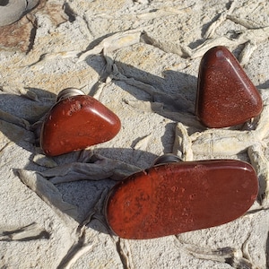 May include: Three red jasper stones with silver accents. The stones are in the shape of a triangle and an oval. The stones are on a light gray textured surface.