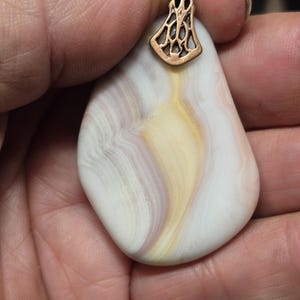 May include: A handcrafted pendant with a teardrop-shaped, white and cream-colored stone, featuring swirling patterns of pale yellow and lavender. It has a decorative, openwork copper-colored metal bail.