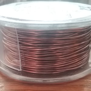 Puede incluir: Una bobina de alambre para manualidades de color cobre oscuro enrollado firmemente alrededor de un carrete cilíndrico de plástico transparente. El carrete cuenta con una pequeña asa lateral y una etiqueta blanca con el número 22 impreso.