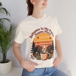 Puede incluir: Una camiseta de manga corta de color crema con un estampado gráfico de una taza de café humeante en un entorno forestal con montañas y una puesta de sol. El texto "Autumn Is My Happy Place" aparece arqueado sobre la ilustración.