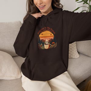 Puede incluir: Una sudadera con capucha de color marrón oscuro que presenta un estampado gráfico de una taza de café humeante, pinos y una puesta de sol. El texto "Autumn Is My Happy Place" aparece arqueado sobre el diseño en una fuente retro.