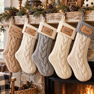 May include: Five cable-knit Christmas stockings in tan, cream, and gray colors. Each stocking features a rectangular leather patch with a name engraved in black script: Mason, Elise, Caleb, Nora, and Liam. The stockings have a ribbed cuff.