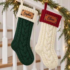 May include: Two cable-knit Christmas stockings hanging side by side. The left stocking is dark green with a cream cuff, and the right is cream with a red cuff. Each features a brown leather tag with the names Sophie and Ethan engraved.