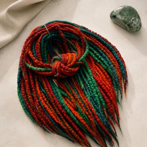 Könnte beinhalten: Ein Set aus synthetischen Dreadlocks in einer lebendigen Mischung aus roten, orangen und blaugrünen Fasern. Die Dreadlocks sind zu einem kreisförmigen Bündel gewickelt und mit einem dünnen Band fixiert, was eine texturierte, gedrehte Optik zeigt.