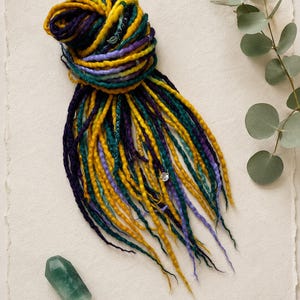 May include: A set of synthetic dreadlocks in shades of mustard yellow, deep purple, teal, and light lavender. The dreadlocks are bundled together at the top and feature small silver-toned metal charms, including a leaf shape and a circular accent.