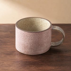 Peut inclure: Une tasse en céramique cylindrique avec un extérieur rose poudré texturé présentant un motif finement quadrillé. L'intérieur est blanc cassé moucheté et l'anse possède une finition texturée assortie à l'aspect sombre et patiné.