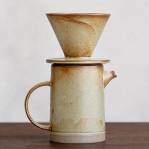 May include: A ceramic pour-over coffee maker set. The set includes a beige-colored carafe with a handle and spout, topped with a matching cone-shaped filter holder. The coffee maker has a rustic, handcrafted appearance.