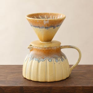 May include: A ceramic coffee dripper and matching carafe set featuring a two-tone glaze. The top dripper is cone-shaped with internal ridges, while the bottom carafe has a ribbed body, a side handle, and a spout, all finished in yellow and brown.