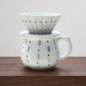 May include: A light blue ceramic coffee dripper with a fluted design and small red dots, resting on a matching light blue ceramic pitcher featuring a pattern of vertical purple brushstrokes and small brown dots, complete with a side handle.
