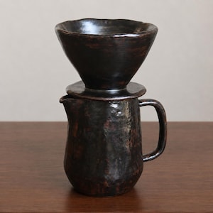 Op de afbeelding: Een donkerbruine keramische koffiefilterset bestaande uit een conische filterkegel met een brede rand die stevig op een bijpassende kan met een zijhandvat en een kleine schenktuit rust, met een getextureerde, onregelmatige oppervlakteafwerking.
