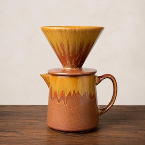 May include: A ceramic coffee pour-over dripper set featuring a conical top piece and a matching pitcher with a handle and spout. The glaze displays a two-tone drip pattern with a warm mustard yellow top transitioning into a deep terracotta brown base.