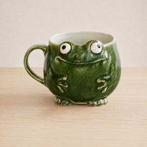 May include: A round, dark green ceramic mug shaped like a frog. The mug features a glossy finish, a curved handle on the left side, and two raised white eyes with black pupils on the top rim. The front displays molded frog arms and feet.