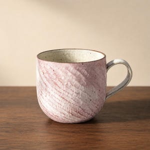 May include: A ceramic mug with a rounded shape, featuring a textured exterior with diagonal carved grooves in a soft pink and white glaze. The interior is a speckled off-white, and the handle is a curved, light-colored ceramic piece attached to the side.