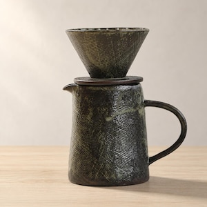 May include: A dark ceramic pour-over coffee maker set. The set includes a textured, cone-shaped filter holder, a lid, and a cylindrical carafe with a handle. The color is primarily dark with green and brown accents. The set is designed for brewing coffee.