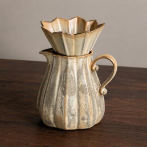 Könnte beinhalten: Ein Keramik-Kaffee-Pour-Over-Set. Das Set beinhaltet eine gerippte, birnenförmige Kanne mit gebogenem Griff und einen blütenförmigen Filterhalter. Das Set ist in Grau- und Beigetönen glasiert.