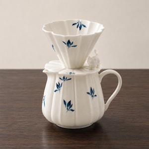 Op de afbeelding: Een witte keramische koffiefilter en kan set met een geribbeld ontwerp en handgeschilderde blauwe bloemmotieven. De kan is voorzien van een klein wit gebeeldhouwd kattenfiguurtje op het deksel en een gebogen handvat om gemakkelijk te schenken.