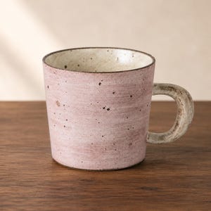 Peut inclure: Une tasse en céramique avec un extérieur rose poudré moucheté et un intérieur blanc cassé lisse. La tasse présente une anse épaisse et incurvée avec une finition grise texturée et vieillie, ainsi qu'un corps cylindrique au bord rustique.