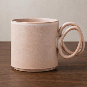 May include: A cylindrical ceramic mug in a muted dusty pink hue with a matte, speckled texture. The mug features a distinctive handle composed of two interconnected, circular loops extending from the side, creating a continuous, open design.