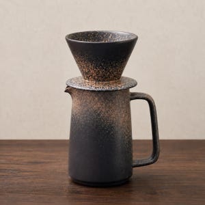 Op de afbeelding: Een keramische koffiefilterset bestaande uit een conische filterhouder op een bijpassende kan. Beide onderdelen hebben een zwarte basiskleur met een textuur van gespikkelde bruine en beige tinten die een verloopseffect op het oppervlak creëren.