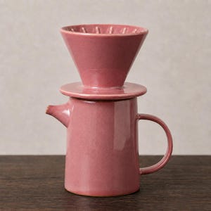 May include: A dusty rose ceramic coffee pour-over dripper set consisting of a conical filter holder with internal ridges resting on top of a matching cylindrical carafe featuring a small spout and a curved side handle with a smooth finish.