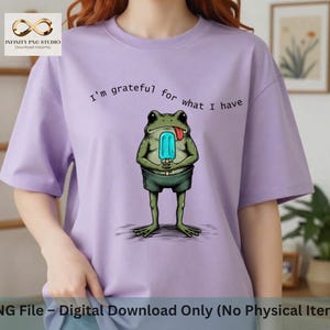 May include: A light purple t-shirt featuring a graphic of a green frog wearing dark green shorts and holding a light blue popsicle. Above the frog, the text reads, I'm grateful for what I have. The shirt has short sleeves and a crew neckline.