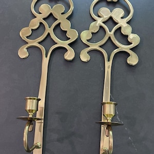 May include: A pair of polished brass wall sconces featuring an elongated vertical stem and a decorative openwork scroll design at the top. Each sconce includes a circular drip plate and a cylindrical candle cup at the base.