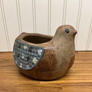 May include: A small, brown, bird-shaped ceramic planter with a textured, light blue wing detail featuring a honeycomb pattern with small white and dark spots. The bird has a simple, dark-colored eye and a smooth, speckled, earthy-toned finish.