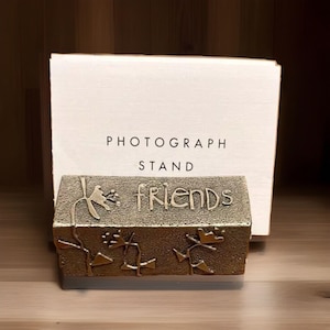 May include: A bronze-colored photograph stand with the word "FRIENDS" in a textured font. The stand has floral designs and is in front of a white box that reads "PHOTOGRAPH STAND".