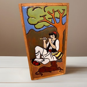 May include: A rectangular ceramic tile with a wooden frame. The tile depicts a man playing a flute under a tree, with a dog lying nearby. The color palette includes brown, green, blue, and white.