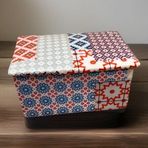 May include: A rectangular storage box featuring a lid and body decorated with a patchwork of colorful geometric patterns. The design includes repeating motifs in shades of red, blue, pink, and white, creating a vibrant and detailed visual display.