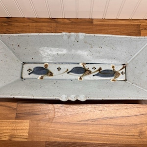 May include: A rectangular ceramic serving tray with a light gray speckled glaze. The tray features two ribbed handles on the ends and a central decorative band painted with three blue leaf-like shapes and small brown floral accents.