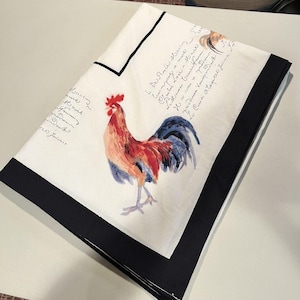 May include: A folded white fabric cloth featuring a central illustration of a rooster with red, orange, and blue plumage. The cloth has a thick black border and is decorated with black cursive script text printed across the white background.