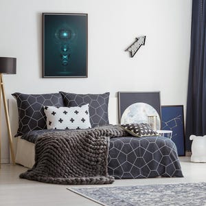 Può includere: Interno di una camera da letto con un letto con un copripiumino e cuscini con motivo geometrico grigio scuro. Una spessa coperta di maglia grigia è sul letto. Un'opera d'arte incorniciata è appesa sopra il letto, e una lampada da terra in legno è a sinistra.