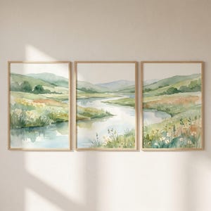 Könnte beinhalten: Ein Set aus drei vertikalen Wandkunstdrucken, die eine durchgehende Aquarell-Landschaft mit sanften grünen Hügeln, einem gewundenen Fluss und Wiesen voller Wildblumen in zarten Grün-, Blau-, Gelb- und Brauntönen zeigen, gerahmt in hellem Holz.