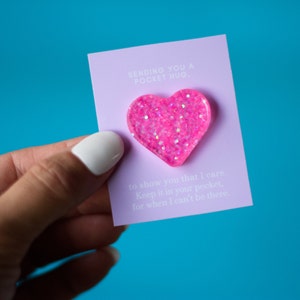 Peut inclure: Épinglette rose en forme de cœur pailleté sur une carte lavande avec le texte "SENDING YOU A POCKET HUG" et un message. L'épinglette est tenue sur un fond bleu vif.
