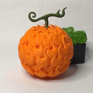 May include: A detailed 3D model of an orange, spherical fruit with a flame-like pattern and a green stem. The fruit is likely a collectible or decorative item. A small black square container with green foliage is in the background.