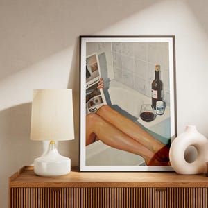Puede incluir: Una impresión artística enmarcada representa a una persona relajándose en una bañera, leyendo un periódico, con una botella de vino y una copa. Una lámpara y un jarrón de cerámica blanca se encuentran en un mueble de madera.