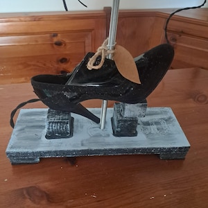 Puede incluir: Una lámpara única con un zapato negro montado sobre una base de madera gris. El zapato está sujeto a un poste de metal, y una etiqueta marrón cuelga del zapato. La base tiene forma rectangular con un acabado pintado.